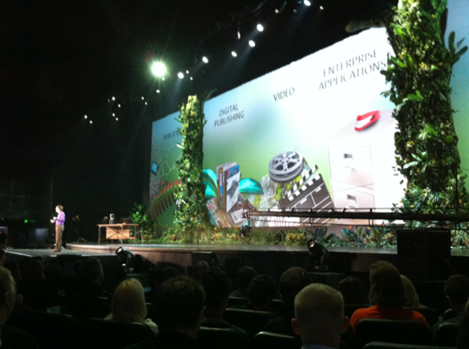A photo of a stage with green theme, while a man in purple shirt gives a lecture on it.