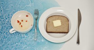 A photo of a plate with a piece of bread and butter, a knife, a fork, a cup with some water and some red seeds. All of them are placed in a blue colored water.