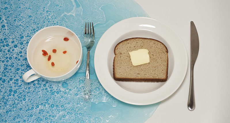 A photo of a plate with a piece of bread and butter, a knife, a fork, a cup with some water and some red seeds. All of them are placed in a blue colored water.