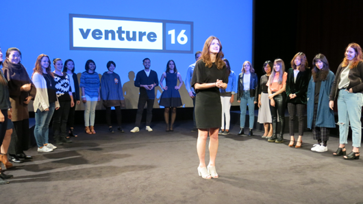 A photo of a group of people standing on a stage around a person, while behind them there is a screen projector with a blue background and the logo VENTURE 16.