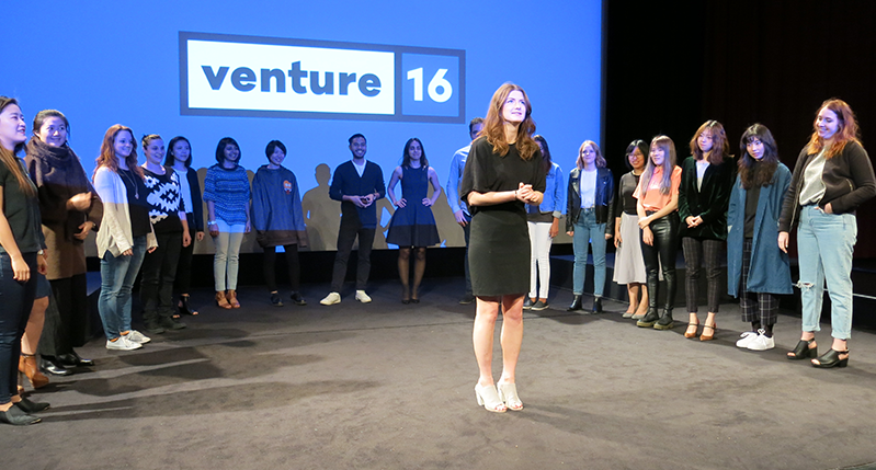 A photo of a group of people standing on a stage around a person, while behind them there is a screen projector with a blue background and the logo VENTURE 16.