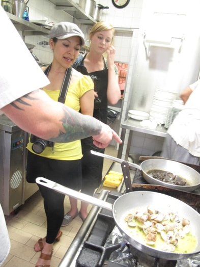 A photo of two woman standing in a kitchen and looking at the food being cooked.