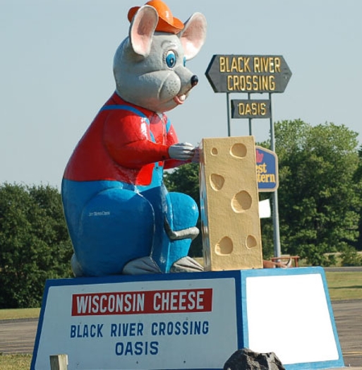 A photo of a Wisconsin Chees mascot that depicts a mouse dressed in a red shirt, a blue union suit and an orange hat while holding a big piece of cheese.