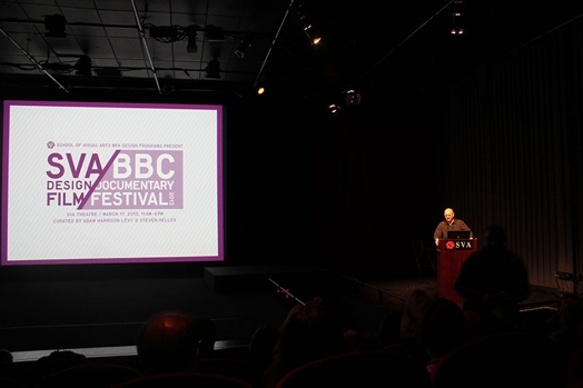 A photo of a man giving a lecture at a stand with SVA logo on it, while near him there is ascreen projector with SVA Design Film BBC Documentary Festival logo.