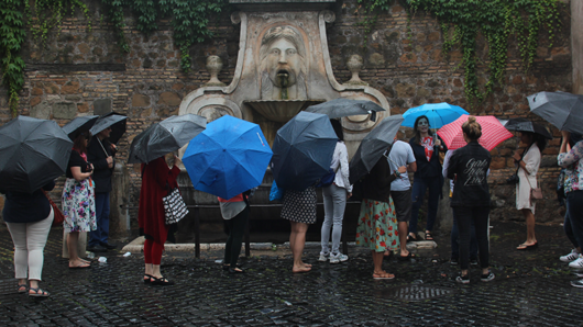 A group of tourists with umbrellas standing near an old roman water fountain with a sculpted head on it.