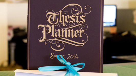 A photo of a book cover with title Thesis Planner made from stylish medieval fonts. Near the cover, placed also on the green table, there is a scroll pf paper tied with a cyan ribbon.