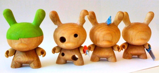 A group of wood sculptures figurines. One of them has green paint on its head. Another one has holes and what looks like an electric drill in its hand.