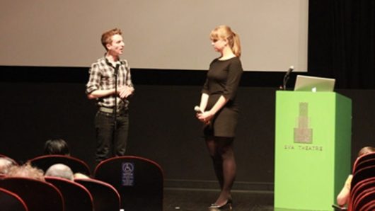 A photo of two people giving a lecture on a stage while sitting near a green stand with a laptop on it.