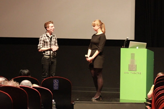 A photo of two people giving a lecture on a stage while sitting near a green stand with a laptop on it.
