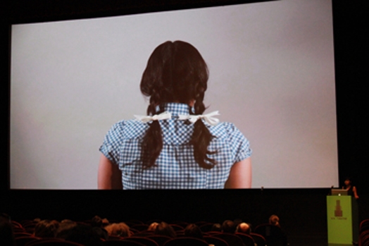 A photo of a screen projection showing a photo of the back of a girl wearing a blue shirt and pony tails.