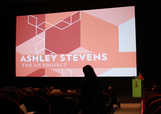 A photo of a screen projection showing of red, orange and white isometric cubes with the title: Ashley Stevens The AH Project.
