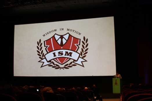A photo of a screen projection showing what looks like a white and red uniform school logo with text: Wisdom In Motion ISM.