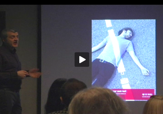 A photo of a person talking on the stage and showing a projection with another person laying on the street with a white stripe over his body.