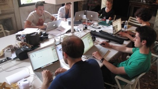 A photo of several students working on laptops at a table while in the middle there is a projector.