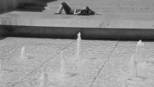 A black and white photo of a fountain spring and a woman which sits next to it and near some stairs.