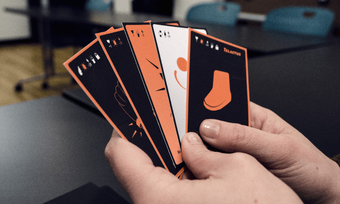 a person holds five playing cards colored in black, orange and white with different illustrations on them
