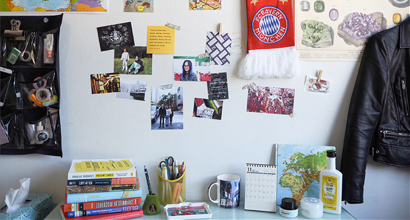 A photo of a desk filled with different items like books, cups, pencil holders, maps, calendars and other bottles. On the wall near the desk there are some photos, banners, maps and other items hanged.