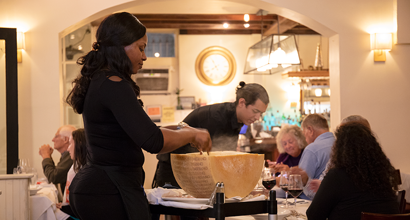 a person preparing a meal in a half-cut parmesan cheese wheel, and in the background are people sitting at the dinner table