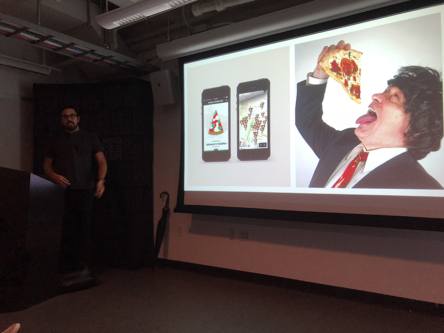 A photo of a man near a screen projector. On it there are a couple of phone templates and a man wearing a suit an d eating pizza.