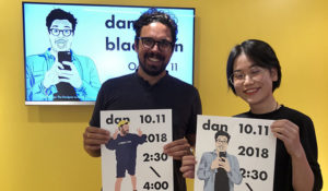A photo of two people holding posters that depict a drawing of a man posing and taking pictures. The text on the posters is: dan.