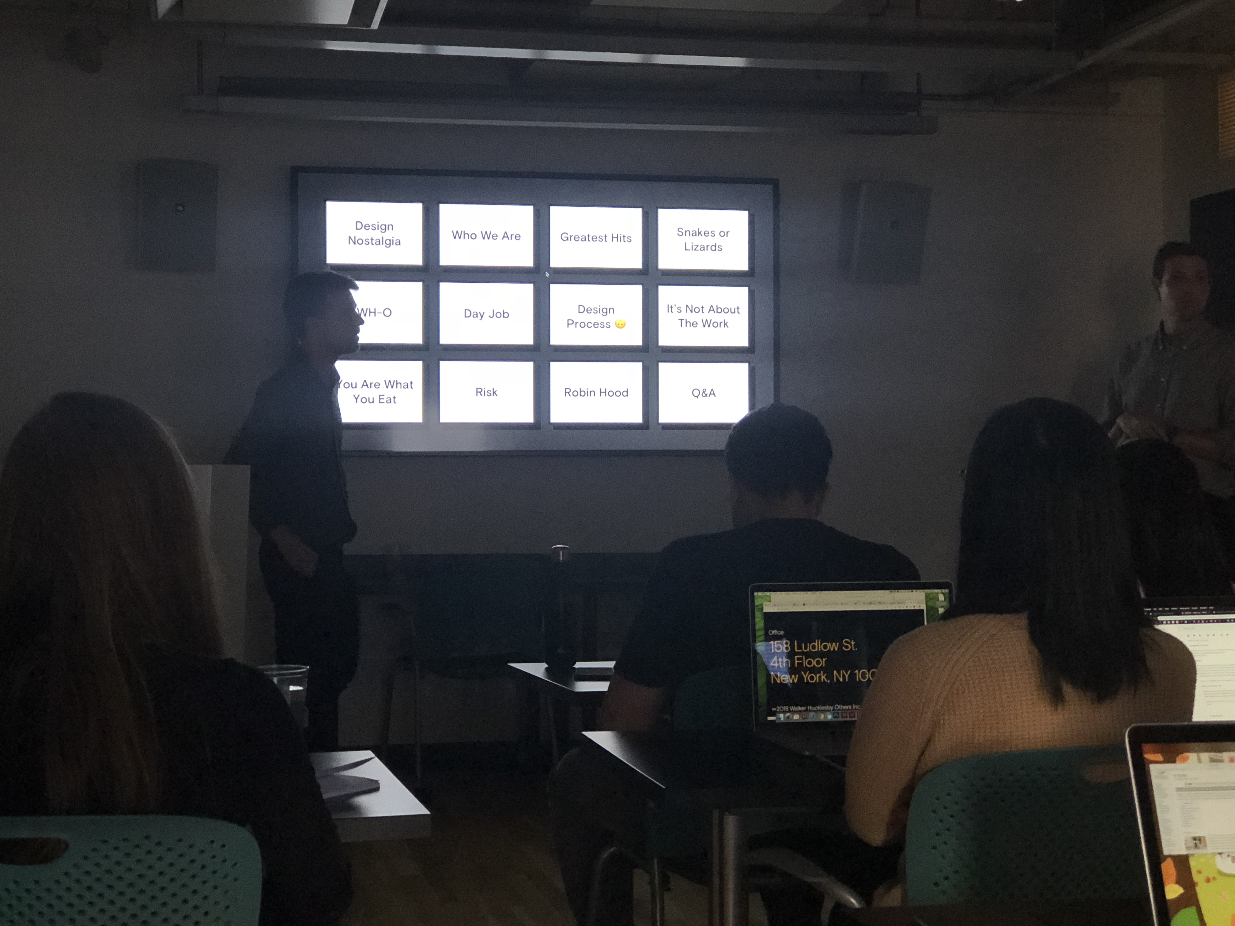 A photo of some students in a classroom listening to a lecture, while a teacher shows them a list of tiles with text on a tv screen.