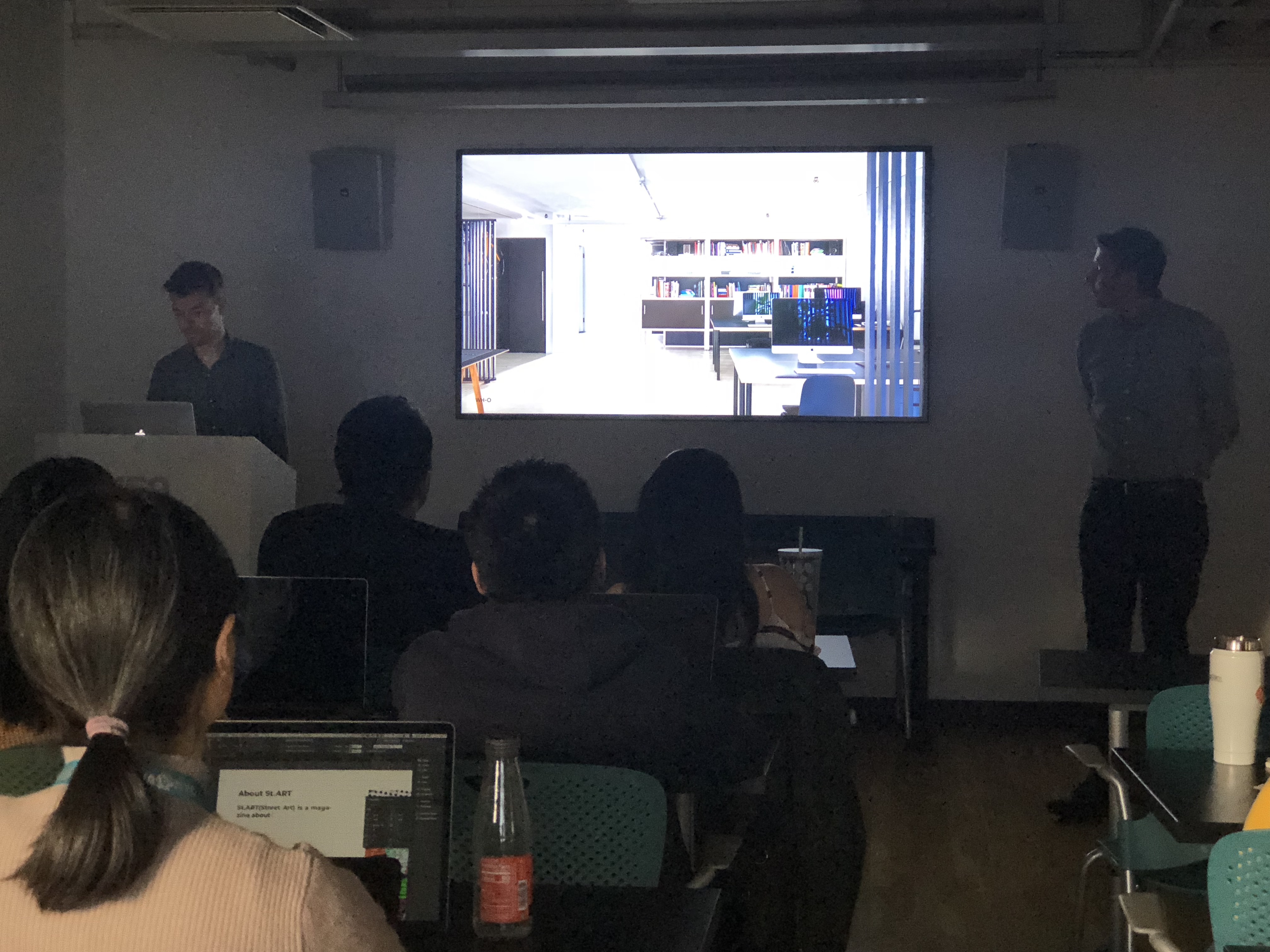 A photo of some students in a classroom listening to a lecture, while a teacher shows them an office room on a tv screen.