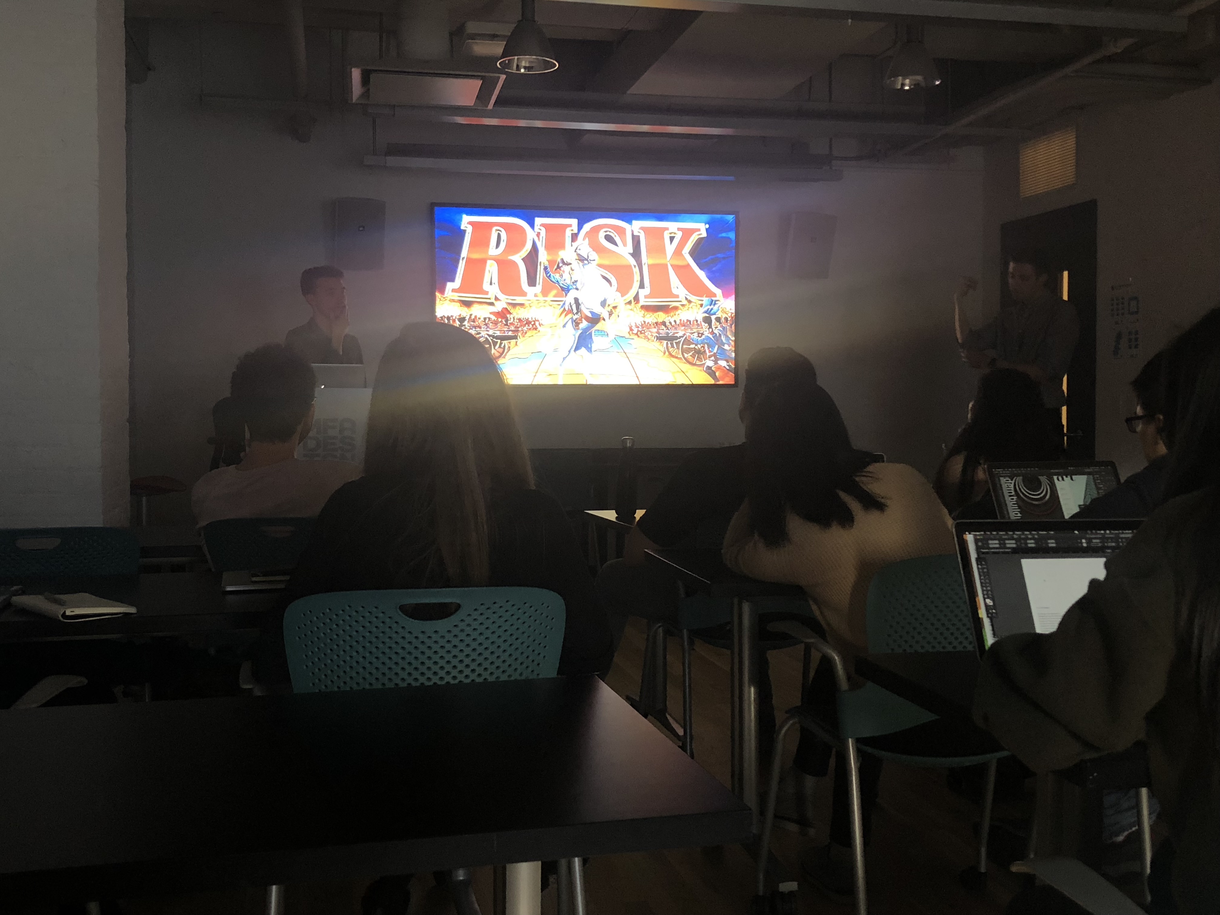 A photo of students sitting in a classroom while a man gives a lecture and shows them a picture of the computer game RISK on a tv screen.