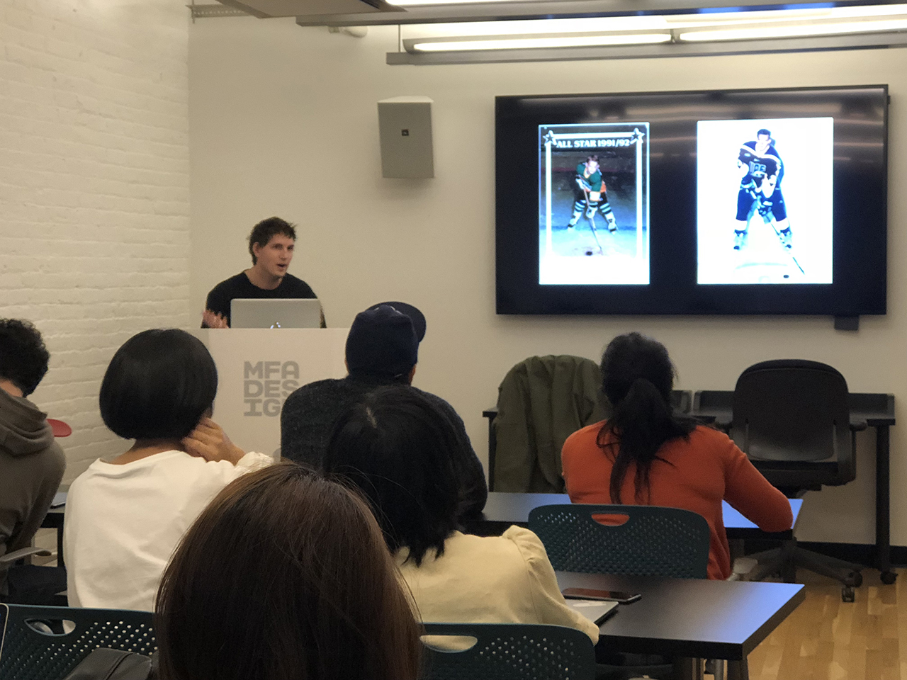 A photo of some students in a classroom, listening to a lecture presented by a man, while watching a tv screen. On the screen there is a picture of a man playing hockey and a card design with the same person.