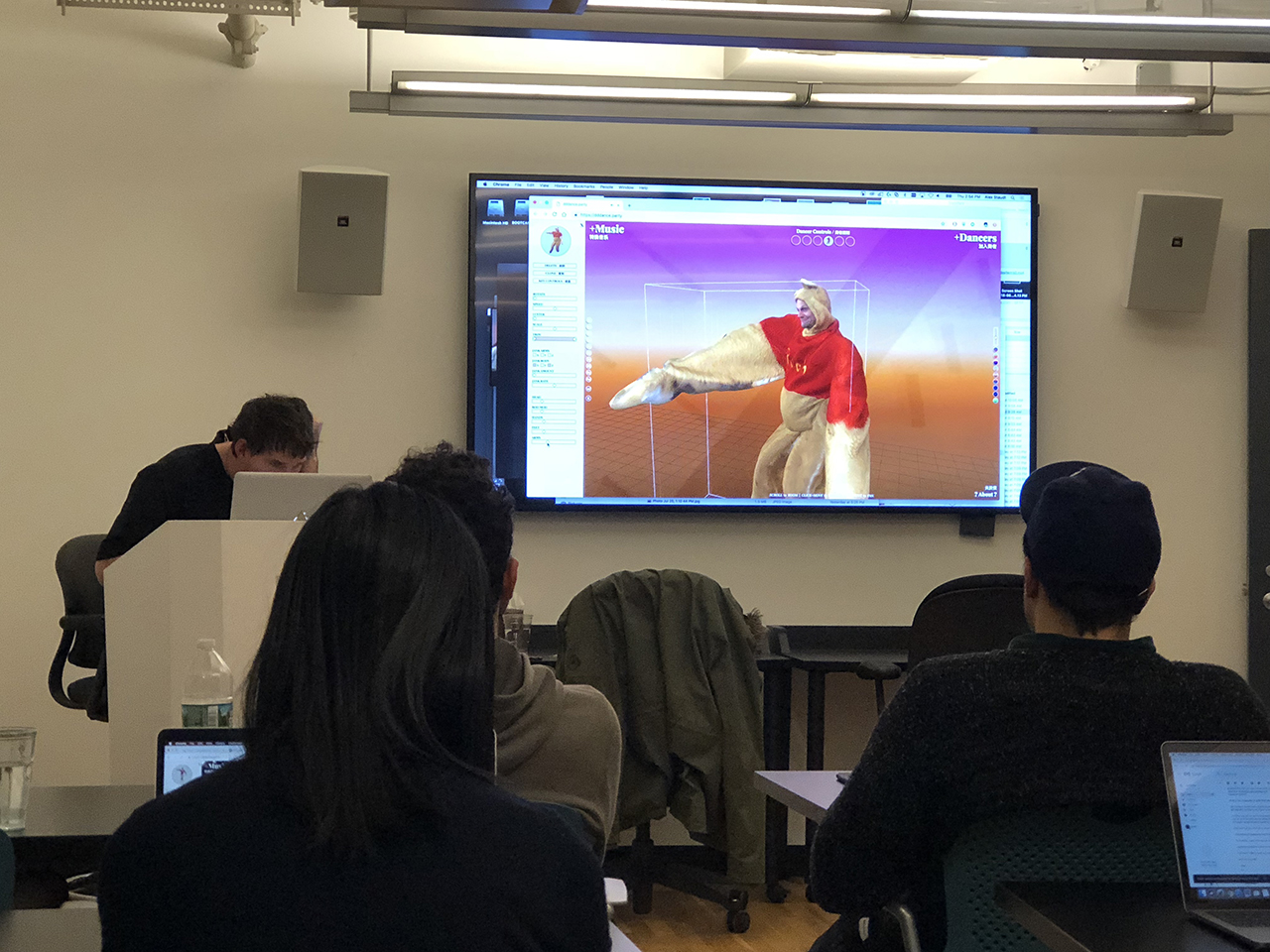 A photo of some students in a classroom, listening to a lecture presented by a man, while watching a tv screen. On the screen there is a 3d rendered image of a distorted humanoid figure.
