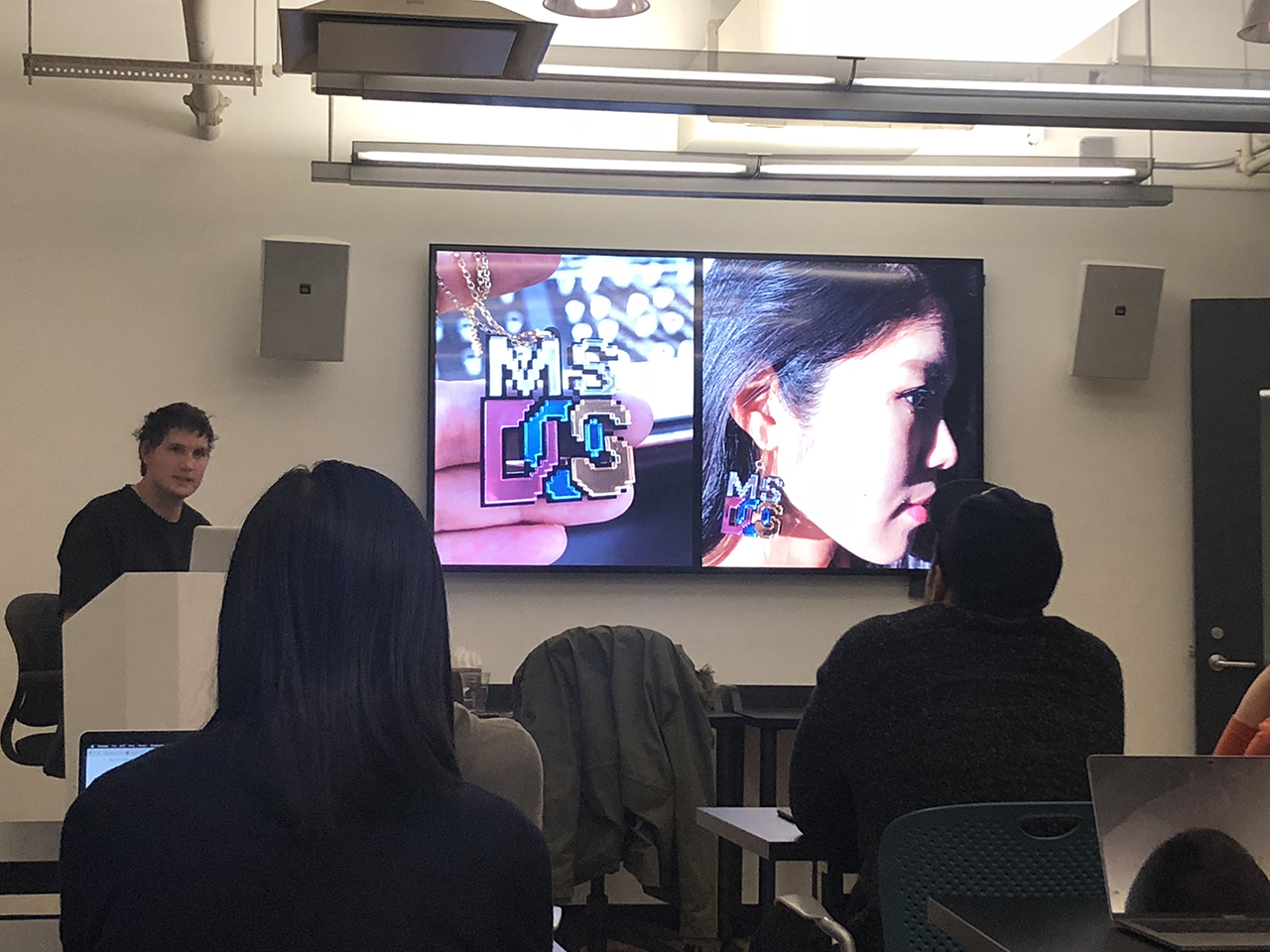 A photo of some students in a classroom, listening to a lecture presented by a man, while watching a tv screen. On the screen there is a photo of a woman wearing some earrings in the form of the MSDOS logo.