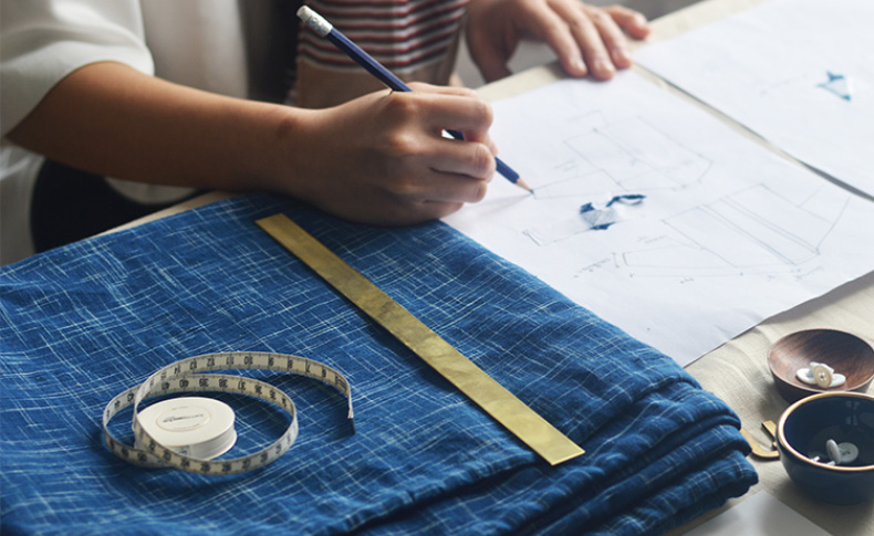 a person designing clothing on a paper, and there are blue fabrics on the table, a measuring scale, and other materials