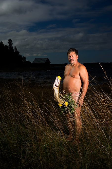 A photo of a naked man sitting in a field, while in front of him there is a big fish with a yellow ball in its mouth. Also the fish ahs some yellow balls and green leaves attached to the tail.