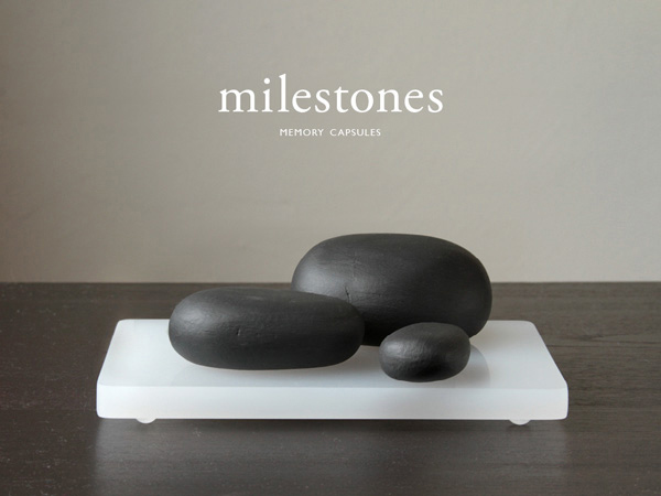 three black stones are installed on a white piece of plastic support on a black table and there is text: milestones, memory capsules