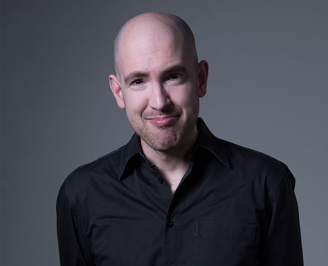 portrait of a bald man smirking in a black shirt and grey background