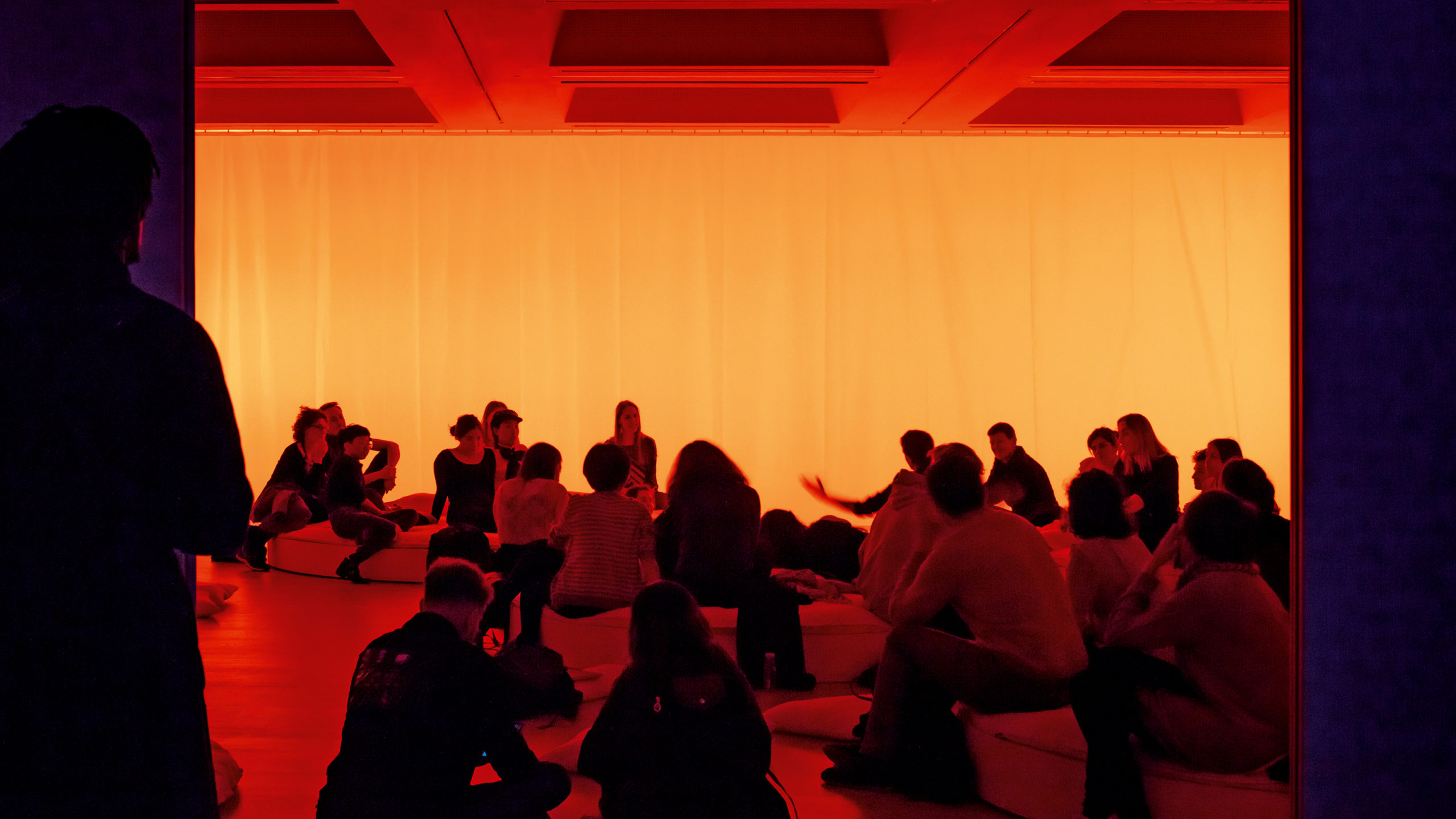 group of people sitting on the floor of an interior space bathed in orange light