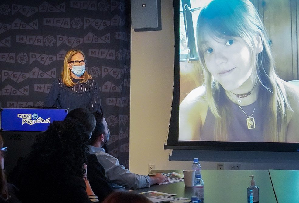 A woman is presenting in front of an audience sitting around the table while she has a picture of a girl projected on the wall on the right side.