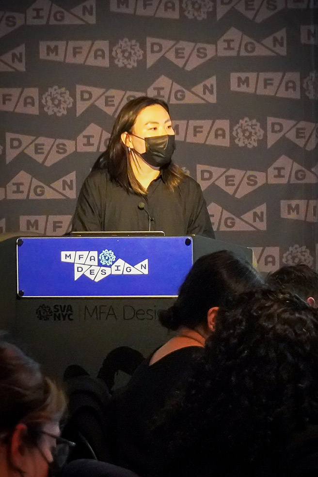 close portrait of a woman wearing a black shirt and a black face mask, with long hair, is giving a speech in front of people
