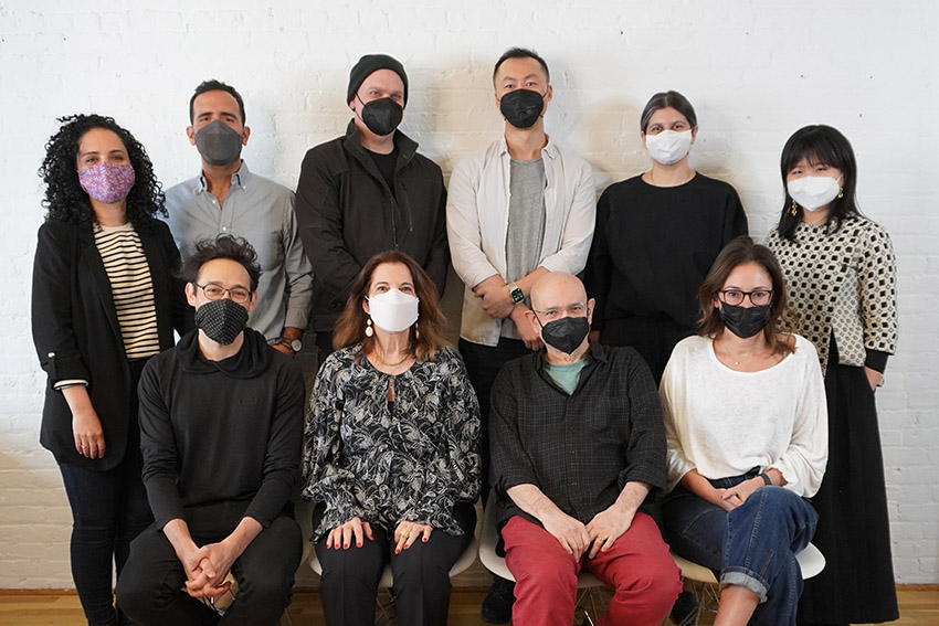 Group portrait with ten people, six standing in the back row and four sitting on chairs in the front row, and they all have face masks on their faces