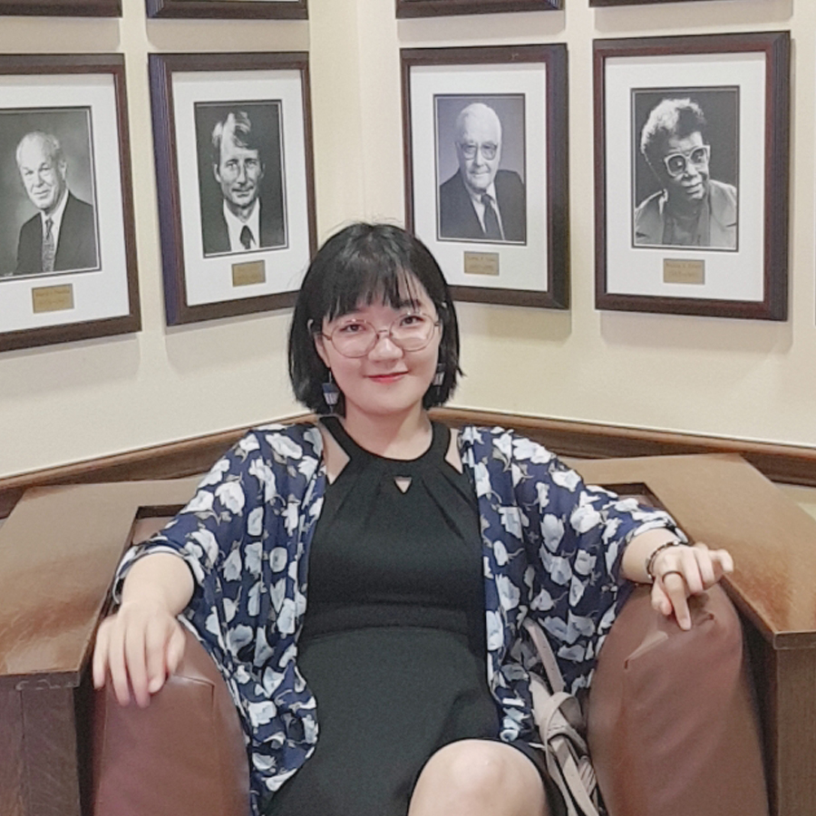 A photo of a woman in a black and blue dress sitting on a couch. Behind here are some blak and white photos of some personalities as well as some golden plated labels.