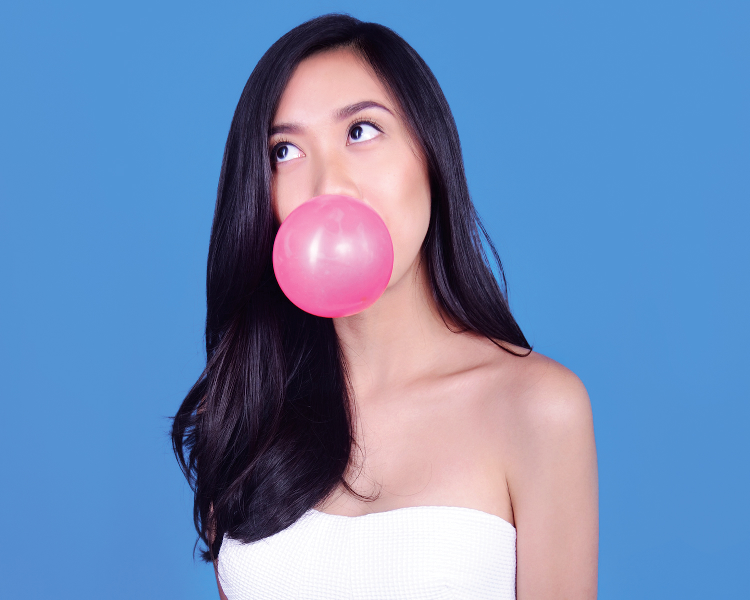 A photo of a woman wearing a white dress and blowing a pink balloon or gum ball.