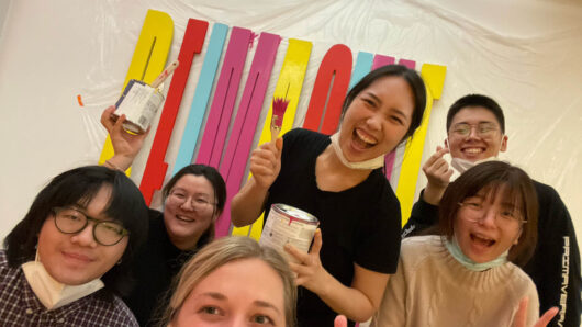 A group of smiling students with paint buckets and brushes in their hands. Also behind them there is a colorful 3d text on a wall that says: REIMAGINE.