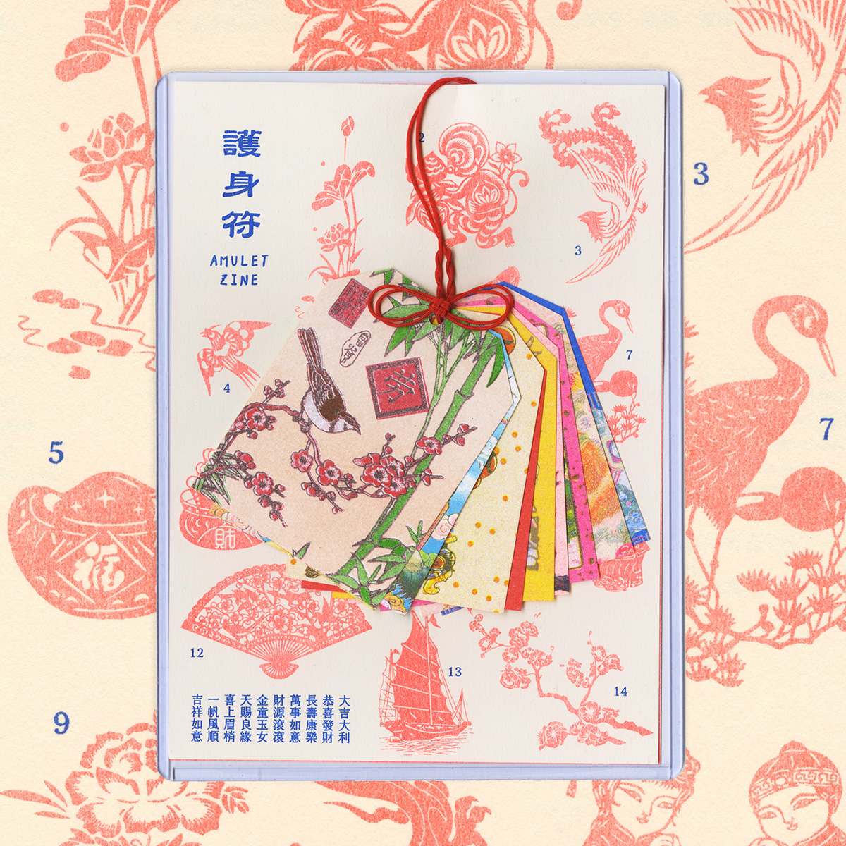 A notebook that has on top of it a set of green, blue, red, yellow, pink and white cloth labels tied together with a red string. On the notebook there are some Asian text and drawings of plants, animals, ships and people.