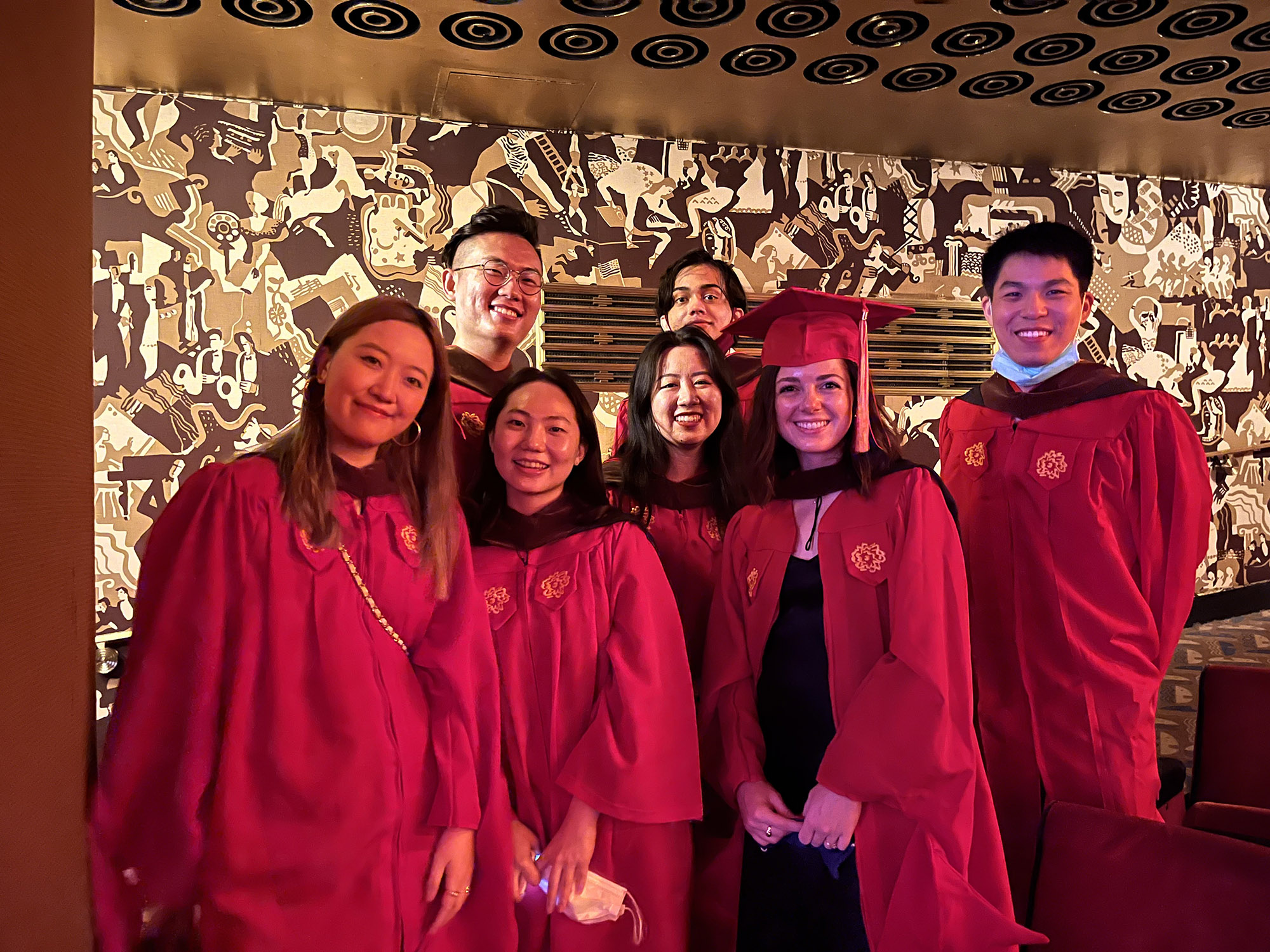 A group of SVA grad students dressed in red robes while standing in a hall with golden plated walls.