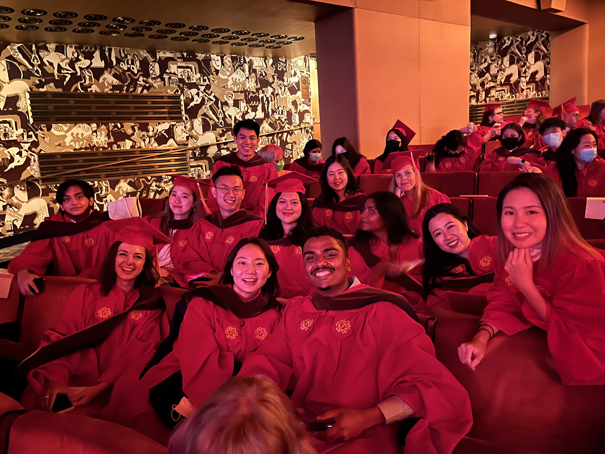 A group of SVA grad students dressed in red robes while sitting in a theatre hall with golden plated walls.