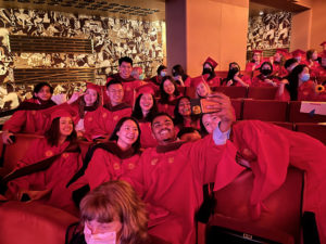 A group of SVA grad students dressed in red robes, and taking selfies while sitting in a theatre hall with golden plated walls.