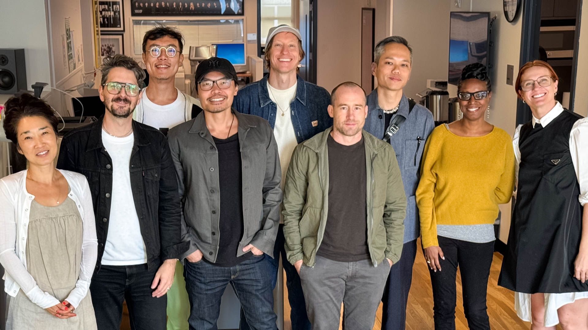 a group portrait of 9 people looking at the camera and smiling. Design studio is the setting.