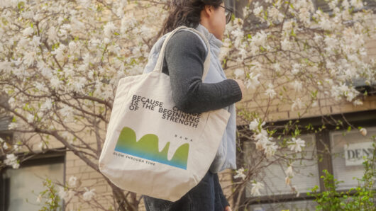 a woman carrying a bag on her shoulder. a flowering tree behind her
