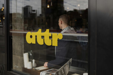 Cafe window with large yellow “attr” logo decal and a person seated inside by the window.