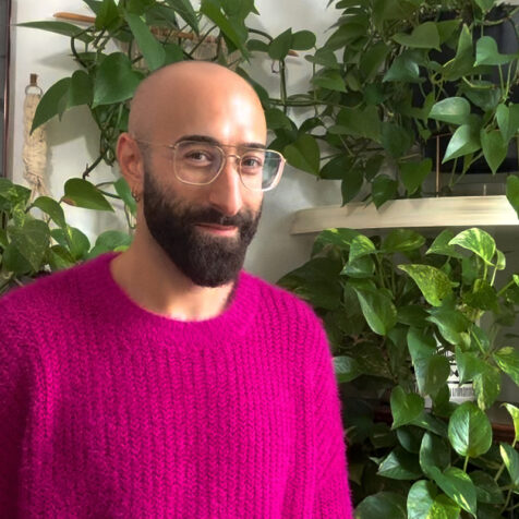 bald man with glasses and a beard looks at the camera. He is wearing a vibrant pink shirt and there are plants behind him.