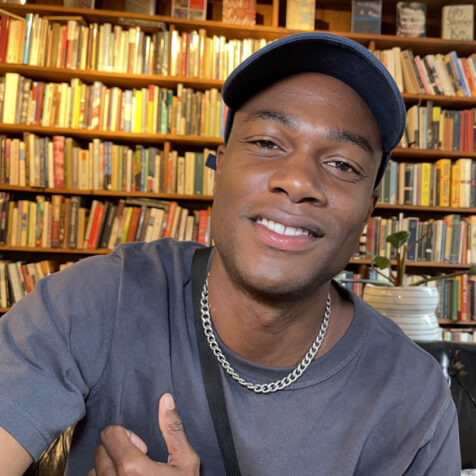 black man with hat looking at camera, books on shelf behind him